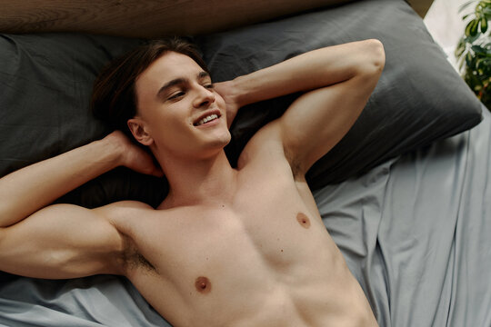top view of smiling and sexy man lying on black pillows with hands behind head and looking away.