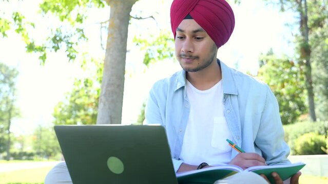 Cheerful indian male student greeting participants of virtual webinar, hindu man in headwrap sits with laptop and waving hello into webcam, has video conference with classmates, taking notes