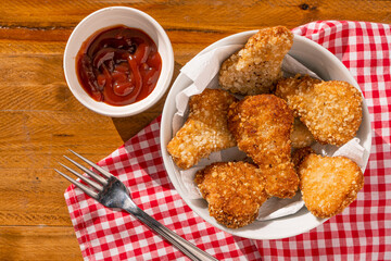 Top view of some fried chicken nuggets in a white bowl accompanied by ketchup sauce on a wooden table.