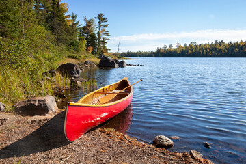 Red wooden canoe on shore of northern Minnesota lake with rocks and pines along the shore