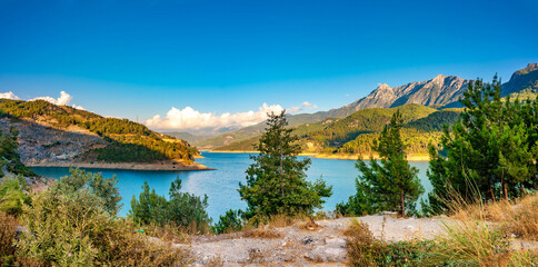 Naklejka premium Beautiful picturesque panorama of a mountainous region with hills covered with forests, a lake and rugged mountains against a blue sky.