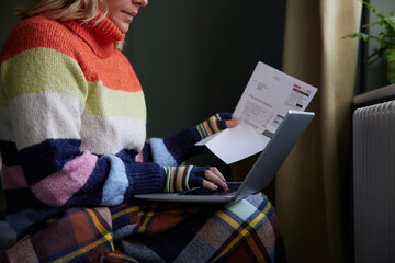 Woman In Gloves With Laptop And Bill Trying To Keep Warm By Radiator During Cost Of Living Energy Crisis