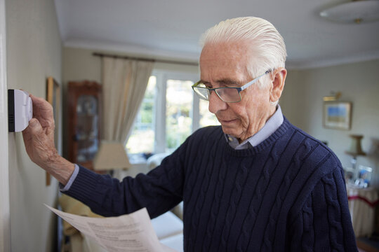 Worried Senior Man With Bill Turning Down Central Heating Thermostat At Home In Energy Crisis