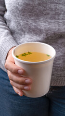 Close-up of Famele hand in grey sweater holding a Chicken broth soup in takeaway cup.