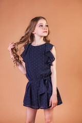 Teen girl princess 10 year old with curly long hair at beige wall background, thinking and looking away. Fashion model in stylish casual blue dress. Fashionable young lady model. Copy text space
