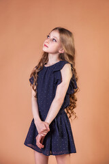 Teenager girl princess 10 year old with curly long hair, standing at beige wall background, looking away. Fashion model in stylish casual blue dress. Fashionable young lady model. Copy text space