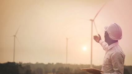 Fototapeta na wymiar African engineer man stands holding tablet front the wind turbines generating electricity power station in sunset. .Concept of sustainability development by alternative energy