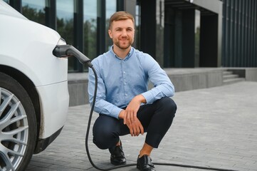 Hansome bearded guy sitting near his new modern electric car and holding plug of the charger, while car is charging at the charging station.