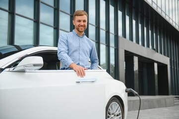 Casual man with smartphone near electric car waiting for the finish of the battery charging process