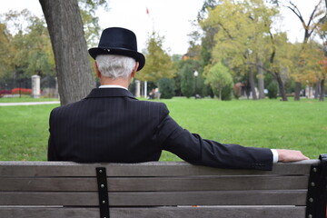 Old man with black hat sitting on a public bench in a park. Grey haired elderly man in elegant suit watching people 