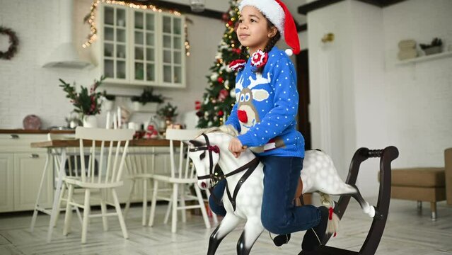 little african american girl in a blue christmas sweater and a red santa hat smiles while sitting on a toy rocking horse against the backdrop of a christmas tree and a festive table.