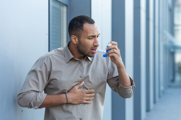 A man outside an office building has a severe asthma attack, a businessman is having difficulty breathing, a worker in a shirt is using an inhaler to make breathing easier, a hispanic man in a casual - obrazy, fototapety, plakaty