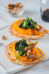 Open sandwich or bruschetta with pumpkin puree, stewed spinach and smoked fish on a white plate against a light concrete background. Antipasti.