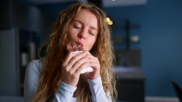 Curly blonde girl overeats unhealthy sweet food. Hungry woman bites into a bar of chocolate and enjoys dessert. Female stuffs her face into candy at kitchen interior. Healthy diet, overweight concept.
