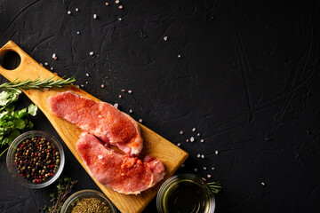 Raw meat steaks on a cutting board. Dark background. Preparation for cooking pork meat. Various spices, seasonings lie nearby. Place for text.