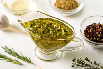 Chimichurri sauce in a gravy bowl on a white background. Various spices lie nearby. Argentinean vegetarian sauce made from olive oil, oregano, parsley.