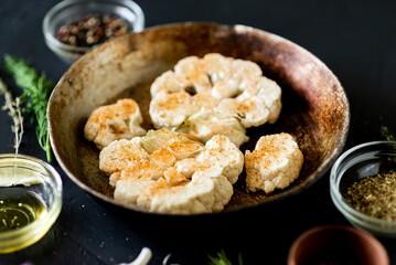 Cauliflower steak cooking. Raw cauliflower sprinkled with spices lies in a frying pan. Olive oil, herbs, various spices nearby. Dark background.