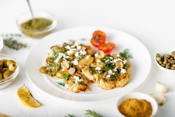 Vegetarian food concept. Cauliflower steak with spices, chimichurri sauce, almond flakes, olives, fried cherry tomatoes and capers on a white plate. White background.