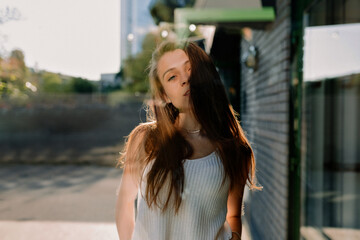 Outdoor photo of stylish young woman with loose hair wearing white t-shirt posing at camera on street inn sunny warm day. Brown-haired woman in spring day. City life concept