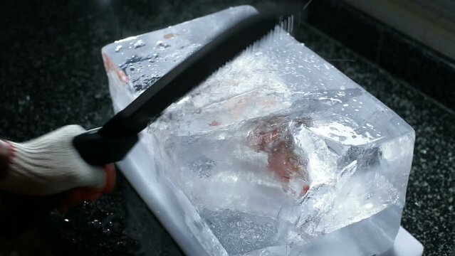 A man uses a machete saw blade to chop and slice a large block of ice with the frozen fish inside the ice cube.