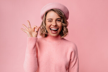 Pretty young caucasian lady winks at camera, shows ok gesture on pink background. Blonde woman with wavy hair wears beret and sweater. Lifestyle, female beauty concept