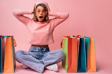 Surprised young caucasian woman holding head with mouth open sitting near packages on pink...