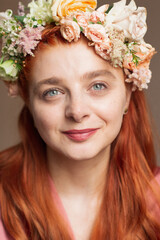 A portrait of a red haired and blue eyed woman, with a flower crown on her head, looking at the camera and smiling 