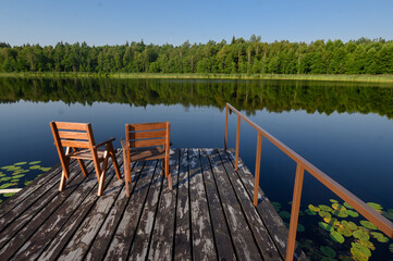 Fototapeta na wymiar Two wooden chairs on a wooden jetty overlooking a calm lake and forest on a sunny day. The concept of relaxation,