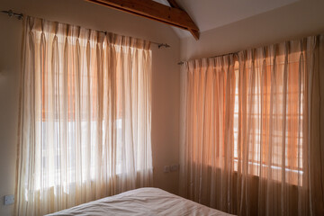 Soft translucent curtains hanging on two windows in a bedroom that has wood beams. There are blinds behind the net type curtains.