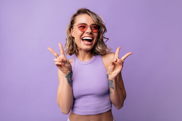 Fototapeta na wymiar Happy young caucasian woman with mood shows victory hand on purple background. Blonde wears crop top and sunglasses in summer. Real emotions concept