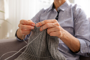 Woman knitting gray wool scarf. Needlework at home. Women’s hobby. Knitting process in women’s hands