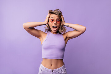 Young caucasian girl with open mouth looks surprised, holds head on purple background. Blonde wears summer crop top and sunglasses. Shock concept