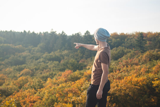 Attractive youthful man in a brown T-shirt and gray cap stands on a rock and points to the leafy trees playing with all the colors of autumn. Wild Sarka, Prague, Czech Republic
