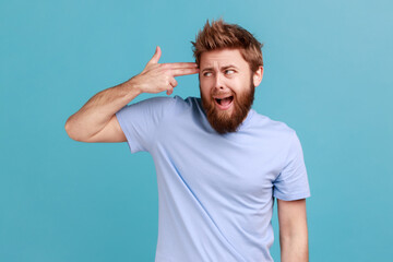 Depression, suicide gesture. Portrait of crazy bearded man standing with finger gun pointed to head, shooting himself with hand pistol. Indoor studio shot isolated on blue background.