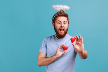 Portrait of excited bearded angelic man with holy nimbus on his head holding two red toy hears,...