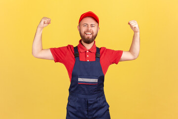 Portrait of proud strong worker man wearing blue uniform and red cap standing with raised arms and...