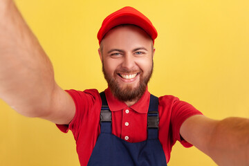Happy excited worker man with wearing red cap and blue uniform taking selfie, looking at camera with toothy smile POV, point of view of photo. Indoor studio shot isolated on yellow background.