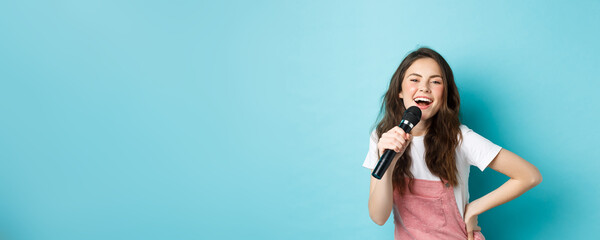 Cheerful young woman singing karaoke, holding microphone and smiling, having fun, standing over...