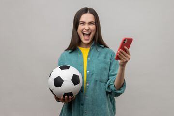 Extremely happy excited woman standing holding soccer ball and using smart phone, betting and winning, wearing casual style jacket. Indoor studio shot isolated on gray background. © khosrork
