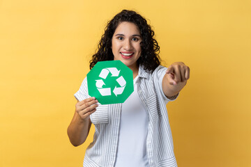 Portrait of satisfied delighted smiling woman with dark wavy hair holding recycling green sign and pointing to camera, saving environment. Indoor studio shot isolated on yellow background.