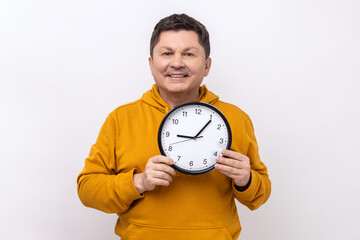 Positive man wearing blue casual shirt showing holding big wall clock, looking at camera with...
