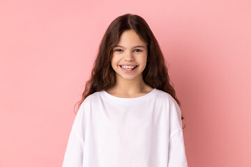 Fototapeta premium Portrait of happy satisfied little girl wearing white T-shirt looking at camera with toothy smile, expressing positive expression. Indoor studio shot isolated on pink background.