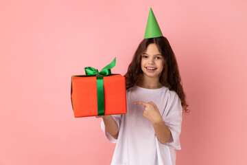 Portrait of little girl wearing white T-shirt and party cone on head pointing at gift box and looking with smile, showing awesome birthday present. Indoor studio shot isolated on pink background.