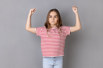 Fototapeta na wymiar Strong dark haired little girl wearing striped T-shirt standing and raising arms, showing her strength and looking at camera with serious expression. Indoor studio shot isolated on gray background.