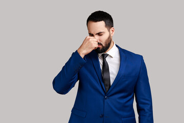 Portrait of bearded man grimacing with disgust, holding breath, pinching nose with fingers to avoid bad smell, awful odor, wearing official style suit. Indoor studio shot isolated on gray background.
