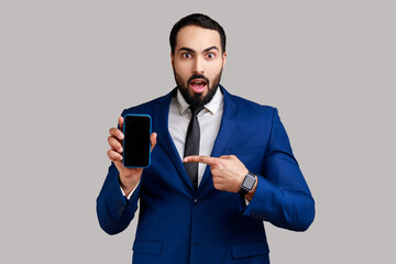 Bearded man pointing finger at smartphone with empty screen, looking at camera with shock, freespace for advertisement, wearing official style suit. Indoor studio shot isolated on gray background.