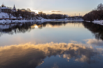 Winter landscape with a river in the old town. Evening, clouds are reflected in the water.