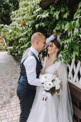 A stylish bearded groom and a beautiful young bride in a white long dress with a diadem on her head stand embracing in the park, growing green plants. Wedding photography, portrait.
