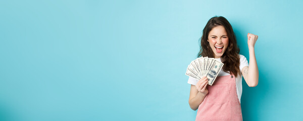 Lucky young woman looks excited, shouting from satisfaction and triumph, winning money, holding...