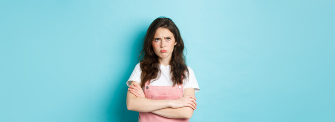 Disappointed sad brunette girl pouting, cross arms on chest and looking at upper left corner unfair...